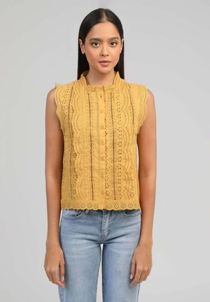 Femme portant un chemisier en dentelle jaune sans manches avec des bords festonnés et un jean bleu clair, debout devant un fond gris uni.