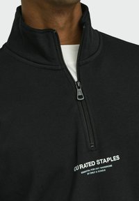 Black pullover with a high collar, half-zip design, and white text branding on the left side. Made of a soft, textured fabric.