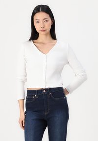 Cardigan cropped a coste bianco con scollatura a V, dotato di chiusura con bottoni e maniche lunghe, abbinato a jeans a vita alta blu scuro.