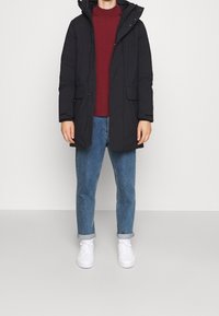 Zwarte gewatteerde jas met een afneembare capuchon, voorzien van zijzakken en een ritssluiting. Draagt over een bordeauxrode top en gecombineerd met blauwe jeans en witte sneakers.