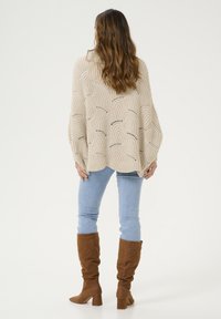 Beige pletený pončo s otevřeným pleteným vzorem, spárovaný se světle modrými džíny a hnědými semišovými vysokými botami, z pohledu zezadu.