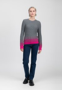 Vrouw staat met een gestructureerde grijze trui met roze accenten, donkere spijkerbroek, zwarte laarzen en zilveren ringen in een lege achtergrond.