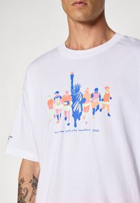 Hvit skjorte med fargerik grafikk av løpere og Frihetsgudinnen, merket "tcs new york city marathon 2025," båret av en person med tatoveringer.