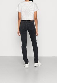 Woman wearing black skinny jeans, white sneakers, and a light gray cropped t-shirt with a subtle tie-dye pattern, standing facing away against a plain wall.