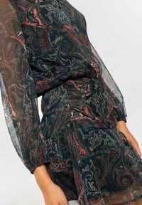 Vestido de patrón oscuro con diseños de paisley y flores en rojo y blanco. Mangas largas transparentes, cintura fruncida y falda en capas.