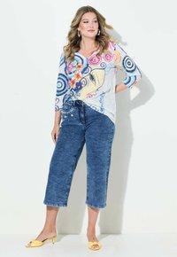 Vícebarevný top s výstřihem do V, pokrytý kruhovými vzory a výšivkou, doplněný o modré zkrácené džíny s květinovými detaily a žluté sandály na podpatku.
