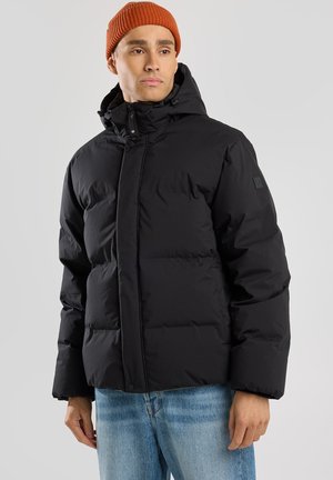 Zwarte gewatteerde jas met hoge kraag, ritssluiting en verstelbare capuchon. Gecombineerd met lichtblauwe denim jeans en een oranje geribbelde beanie.