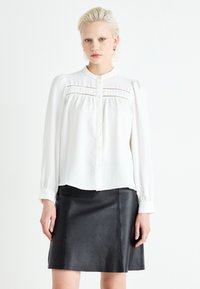 Bílá halenka s dlouhými rukávy, detaily s texturovanými panely a malým límečkem, kombinovaná s černou koženou mini sukní. Hladká tkanina, minimalistický design.