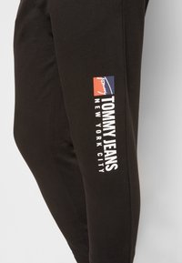 Svarta bomulls-joggers med en sidologga i röd, vit och blå som visar "TOMMY JEANS NEW YORK CITY" i fet typografi.