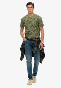 Camiseta verde con un patrón de bambú negro, jeans azules y una camisa de cuadros atada a la cintura. Zapatillas de color claro completan el atuendo.