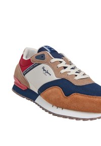 Sneaker multicolori con suede marrone, tela blu navy, accenti rossi e una suola intermedia bianca. Include un design con lace-up e branding.