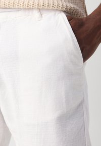 Pantaloni bianchi texture con una vestibilità rilassata, caratterizzati da tasche laterali e una cintura liscia. Il materiale sembra leggero e traspirante.