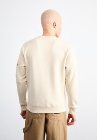 Bézs pulóver bordázott szegéllyel és mandzsettával, hátul téglalap alakú folttal. Barna cargo nadrággal viselve, laza, kényelmes fazont mutatva.