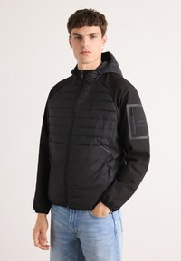 Schwarze Pufferjacke mit Kapuze, bestehend aus einer Kombination aus gesteppten und glatten Stoffen, mit Reißverschlusstaschen an den Seiten und einer Tasche am Ärmel.