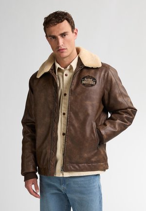 Chaqueta de cuero marrón con cuello de borrego, presenta un parche con el logo, puños acanalados y bolsillos con cremallera. Diseño casual con un acabado ligeramente desgastado.