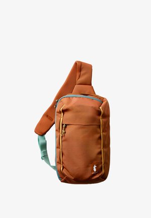 Mochila bandolera naranja óxido con correa acolchada, bolsillo delantero con cremallera y detalles laterales en color teal, con una pequeña etiqueta blanca con logo de un animal.