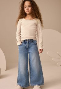 Helle beige Langarmbluse kombiniert mit übergroßen blauen Denim-Jeans mit hoher Taille, weitem Bein und abgenutzten Akzenten.