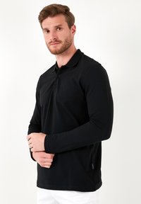 Čierna polo košieľka s dlhým rukávom vyrobená z hladkej látky, s golierom, dvoma gombíkmi a detailom postranného štítku. Minimalistický dizajn.