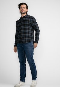 Zwart en blauw geruite flanellen shirt met knopen aan de voorkant, gedragen met slim-fit blauwe jeans en witte sneakers, staande tegen een witte achtergrond.