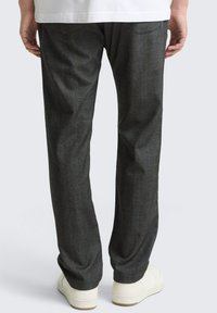 Mann, der dunkelgraue gerade geschnittene Hose und weiße Turnschuhe trägt, von hinten vor hellem, einfarbigem Hintergrund gezeigt.