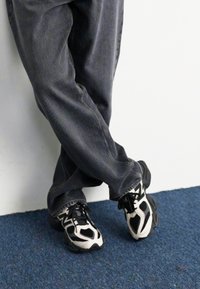 Black and white sneakers with a patterned upper, worn with gray denim jeans, standing on blue carpet against a white wall.