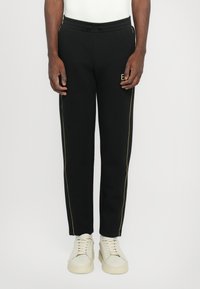 Pantalons de sport noirs avec des rayures dorées sur les côtés, une taille élastique avec cordon de serrage et un petit logo. Assortis à des baskets ivoire.