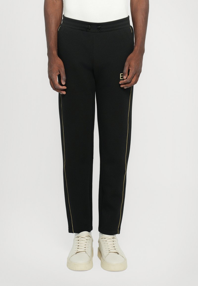 Pantalons de sport noirs avec des rayures dorées sur les côtés, une taille élastique avec cordon de serrage et un petit logo. Assortis à des baskets ivoire.
