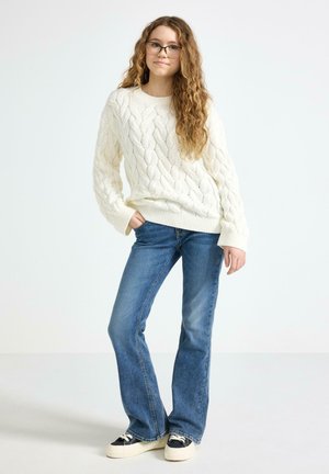 Nuori henkilö, jolla on pitkät kiharat hiukset ja silmälasit, pukeutuneena valkoiseen palmikkoneulepaitaan, sinisiin farkkuihin ja mustavalkoisiin tennareihin, seisoo yhdellä kädellä taskussa.