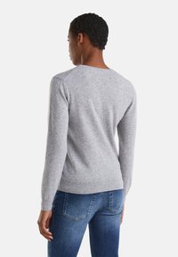 Osoba z krótkimi, kręconymi włosami ubrana w jasnoszary sweter z długimi rękawami i niebieskie dżinsy, odwrócona plecami na tle białej ściany.