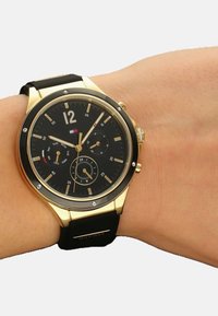 Montre analogique dorée avec un cadran noir, un bracelet en caoutchouc noir et des accents en diamant. Les caractéristiques comprennent des sous-cadrans et un indicateur de date.
