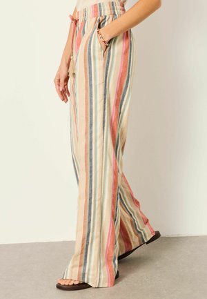 Person wearing loose, wide-leg pants with vertical beige, orange, pink, blue, and white stripes, with one hand in pocket and sandals.