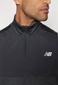 Veste légère zippée noire avec col montant, dotée d'une partie supérieure texturée et d'une section inférieure lisse, ornée d'un logo réfléchissant sur la poitrine.
