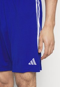 Pantalones cortos deportivos azules de tejido ligero, con rayas blancas a los lados y un logo plateado de Adidas en el dobladillo inferior.