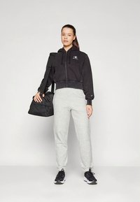 Juodas trumpintas džemperis su logotipu, derinamas su šviesiai pilkomis laisvalaikio kelnėmis ir juodais sportbačiais. Per vieną petį permesta juoda sportinė rankinė.