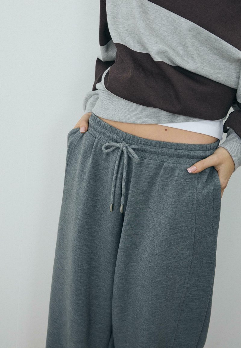 Person wearing loose grey sweatpants with drawstring and striped grey and dark brown long-sleeve top, hands resting in pockets.