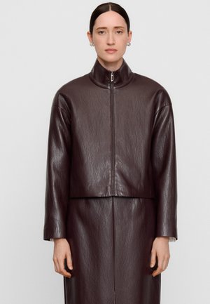 Femme portant une veste en cuir marron foncé avec un col montant zippé, associée à une jupe en cuir assortie, debout devant un fond uni.