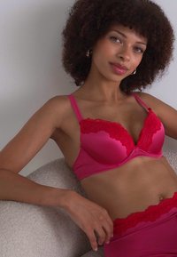 Soutien-gorge en dentelle rose avec un tissu lisse, présentant des bretelles réglables et un design balconnet, complété par un bas assorti en rose.