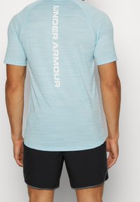Camiseta deportiva de color azul claro con un acabado texturizado y logo "UNDER ARMOUR" en blanco en la parte posterior; combinada con pantalones cortos negros con acentos grises.