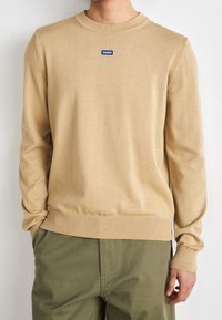 Beige neulepaita, jossa on pyöreä kaula-aukko. Rinnassa sininen "HUGO"-logo ja ribbaatut hihansuut sekä helma.