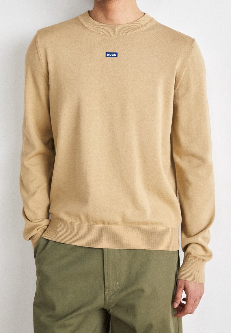 Beige neulepaita, jossa on pyöreä kaula-aukko. Rinnassa sininen "HUGO"-logo ja ribbaatut hihansuut sekä helma.