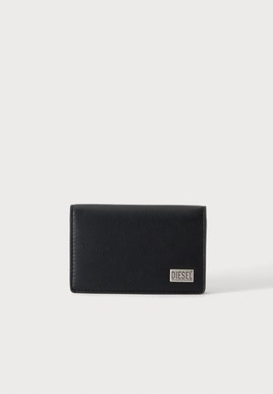 Portefeuille en cuir noir avec une petite plaque argentée portant le logo Diesel dans le coin inférieur droit, posé sur un fond clair uni.