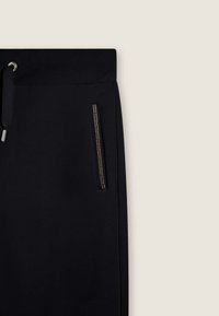 Pantalon de jogging noir avec taille à cordon de serrage, comportant une poche zippée avec des accents métalliques sur le côté. Texture de tissu douce et lisse.