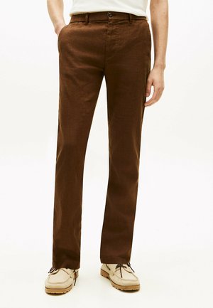 Mann trägt braune, gerade geschnittene Hose mit Vordertaschen, kombiniert mit hellbeigen Schnürschuhen vor einem schlichten weißen Hintergrund.