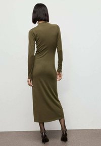 Abito verde oliva a maniche lunghe con silhouette aderente, che arriva a metà polpaccio. Tessuto liscio e collo rotondo discreto, abbinato a scarpe con tacco alto.