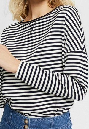 Kobieta ubrana w czarno-biały, poziomo paskowany długi rękaw, włożony do wysokich, niebieskich dżinsów z guzikami.