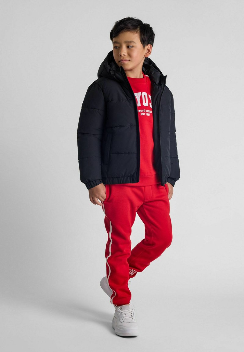 Svart puffjacka med huva, röd sweatshirt med vit text, röda sweatpants med sidostripes, och vita sneakers.