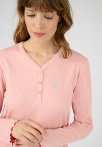 Maglietta a maniche lunghe, rosa a coste con scollatura a onda, tre bottoni e ricamo di un cuore argentato sul petto. Materiale in cotone morbido.