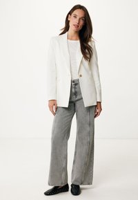 Bílý přizpůsobený blejzr s dlouhými rukávy přes světle šedé tričko, doplněný o kalhoty s vysokým pasem a širokými nohavicemi v šedé barvě a černé slip-on boty s cvočky.