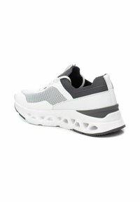Zapatilla deportiva en blanco y gris con parte superior de malla, cuello acolchado y suela gruesa y acolchada que presenta grandes orificios para mayor flexibilidad.