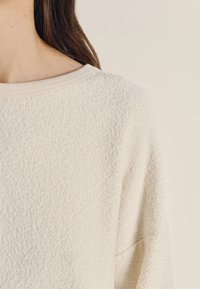 Tekstūruotas, kreminės spalvos megztinis su plačiu iškirptu ir banguotomis rankogaliais. Jame yra minkštas, šiek tiek pakilęs audinio paviršius, suteikiantis šilumą ir komfortą.